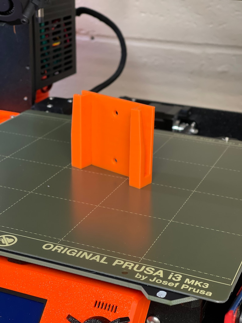 Op de afbeelding: Een oranje 3D-geprint onderdeel met twee gaten op een grijs, getextureerd oppervlak. Het oppervlak heeft de tekst "ORIGINAL PRUSA i3 MK3 by Josef Prusa".