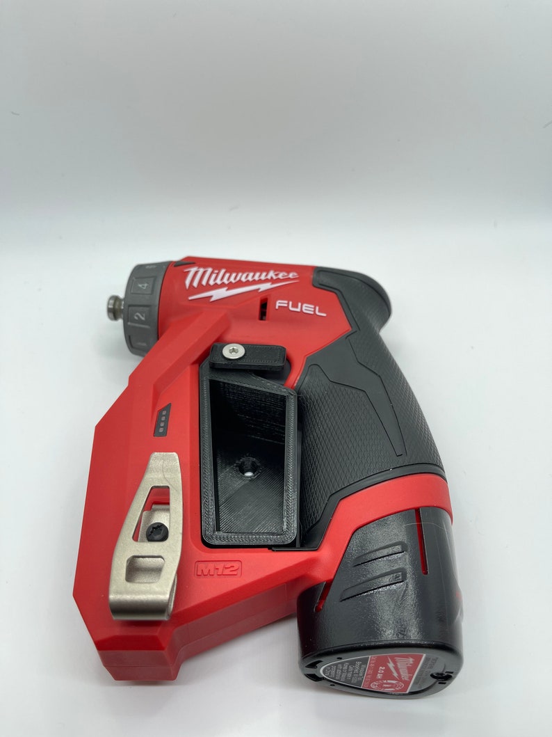 Puede incluir: Un taladro inal&aacute;mbrico Milwaukee FUEL M12 rojo y negro con un clip para cintur&oacute;n. El taladro tiene un agarre de goma negro y un gatillo plateado.