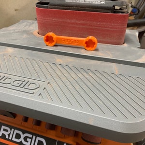 Puede incluir: Una lijadora de banda Ridgid gris y naranja con una banda de lijado roja. La lijadora tiene una llave con el logotipo de Ridgid.