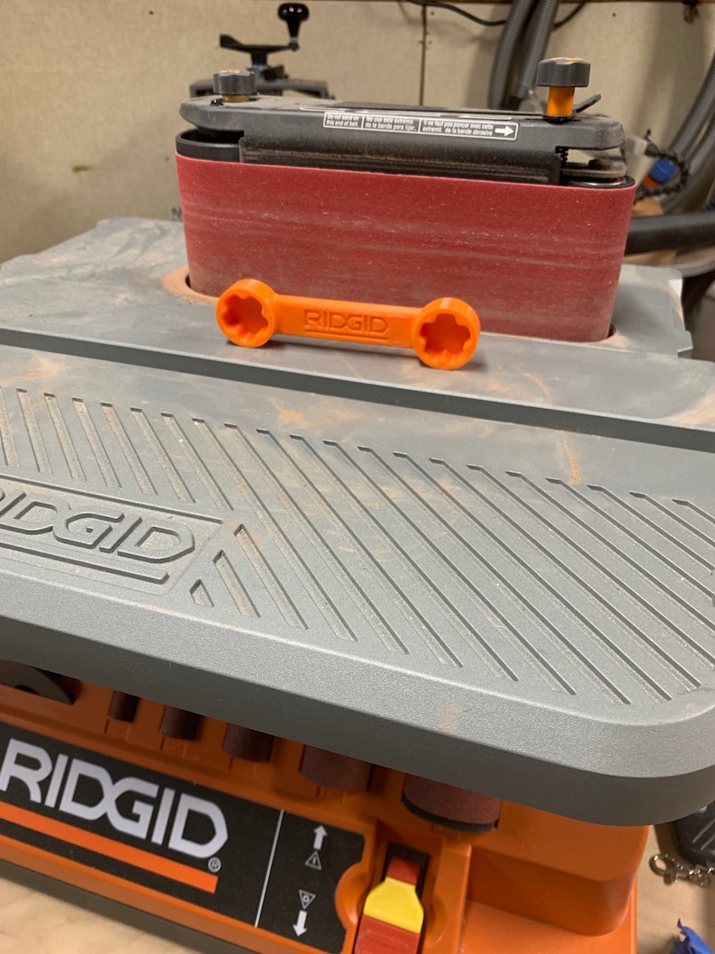 Puede incluir: Una lijadora de banda Ridgid gris y naranja con una banda de lijado roja. La lijadora tiene una llave con el logotipo de Ridgid.