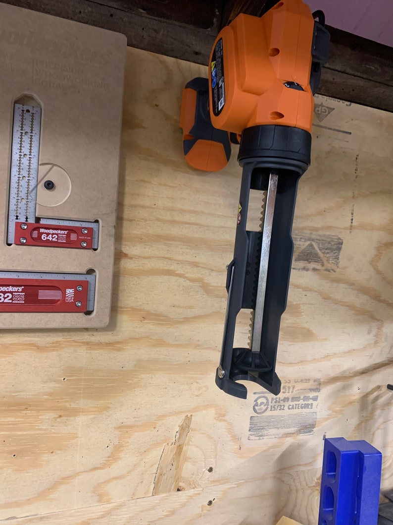 Op de afbeelding: Een oranje en zwarte draadloze kitspuit met een metalen spuitmond. De spuit hangt aan een muur met een houten lambrisering op de achtergrond.