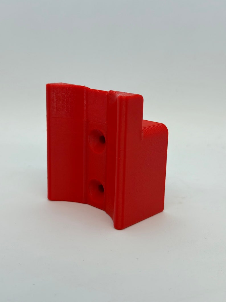 Op de afbeelding: Een rood plastic 3D-geprint onderdeel met twee gaten. Het onderdeel is gevormd als een beugel met een gebogen gedeelte.