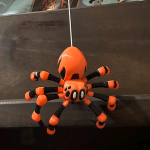 May include: A bright orange and black spider toy with a string attached to its back. The spider has large, white eyes and black spots on its body.