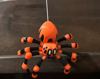 Puede incluir: Un juguete de araña de color naranja brillante y negro con una cuerda atada a su espalda. La araña tiene ojos grandes y blancos y manchas negras en su cuerpo.