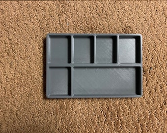 Può includere: Un organizer rettangolare grigio con più scomparti. L'organizer ha una serie di piccole sezioni quadrate e rettangolari, ideali per riporre piccoli oggetti. L'articolo è realizzato in un solido materiale grigio opaco.