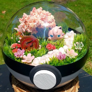 May include: A glass dome with a black and white Pokeball base, containing a miniature pink and white Pokemon figurine, a pink cherry blossom tree, and white rocks. The dome is filled with green grass and pink flowers.