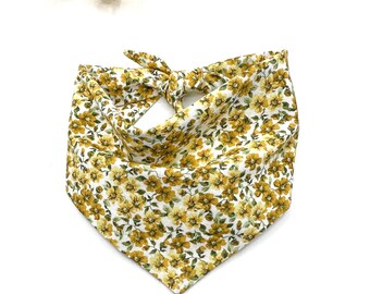 May include: A triangular pet bandana featuring a repeating pattern of small yellow flowers with green leaves on a white background. The fabric is tied at the top with a knot, creating a folded, pointed shape that hangs downward in a classic style.
