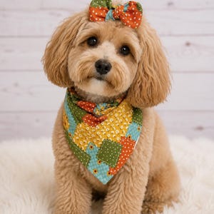 May include: A dog wearing a triangular bandana and a matching hair bow. The bandana and bow feature a patchwork pattern with squares in shades of green, orange, and yellow, decorated with small white floral and polka dot prints throughout.