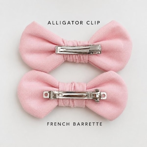 May include: Two light pink fabric bows. The top bow features a silver metal alligator clip, and the bottom bow features a silver metal French barrette. The text ALLIGATOR CLIP is above the top bow, and FRENCH BARRETTE is below the bottom bow.
