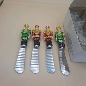May include: Four metal cheese spreaders with handles shaped like nutcracker figures. Two figures wear green jackets with gold crowns, and two wear red jackets with gold hats. Each spreader has a flat, wide, silver-colored stainless steel blade.