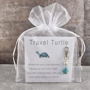 Könnte beinhalten: Ein weißer Organza-Geschenkbeutel mit einer Karte mit der Aufschrift "Travel Turtle" und einer Schildkröten-Grafik. Ein silberner Schlüsselanhänger mit einem Schildkröten-Anhänger und Perlen ist befestigt. Die Karte enthält ein Gedicht über das Reisen.