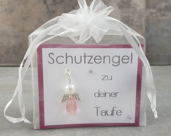 Puede incluir: Una bolsa de organza blanca con un amuleto de ángel rosa y blanco. El amuleto tiene una perla y alas de cristal rosa. La bolsa tiene una etiqueta que dice "Schutzengel zu deiner Taufe".