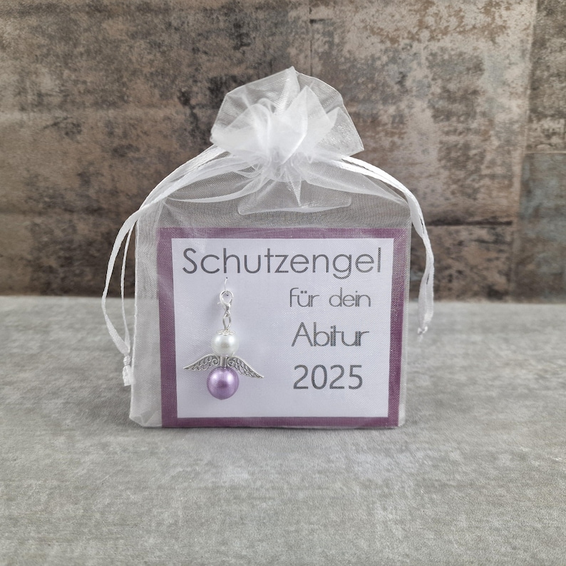 May include: A small white organza bag with a silver angel charm and a purple bead. The bag has a card attached to it that says "Schutzengel f&uuml;r dein Abitur 2025".
