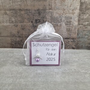 May include: A small white organza bag with a purple card inside. The card has the text "Schutzengel f&uuml;r dein Abitur 2025" and a silver angel charm with a purple bead.