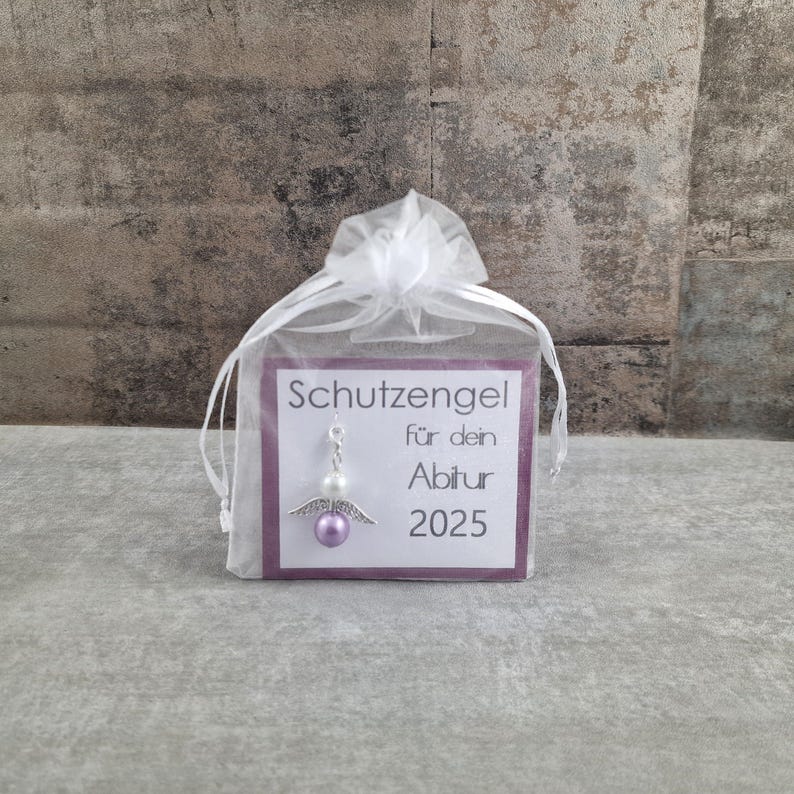 May include: A small white organza bag with a purple card inside. The card has the text "Schutzengel f&uuml;r dein Abitur 2025" and a silver angel charm with a purple bead.