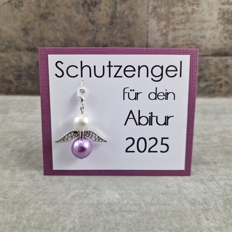 May include: A purple card with the text "Schutzengel f&uuml;r dein Abitur 2025" and a silver angel charm with white and purple beads.
