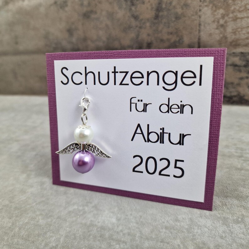 May include: A purple and white angel charm with wings, attached to a card that reads "Schutzengel f&uuml;r dein Abitur 2025".