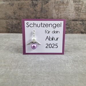 May include: A purple card with white text that reads "Schutzengel f&uuml;r dein Abitur 2025". A small silver angel charm with white and purple beads hangs from the card.