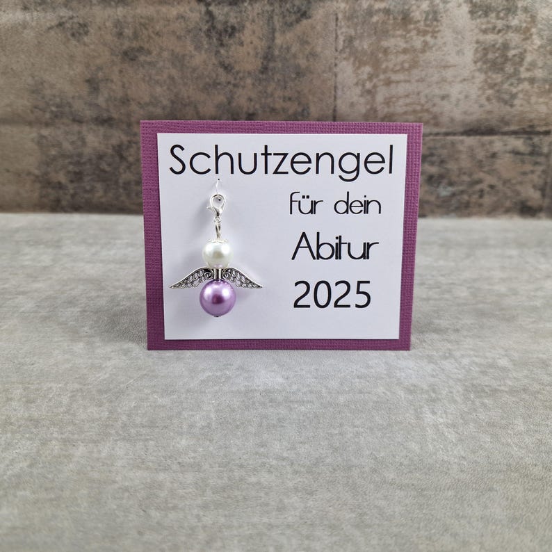 May include: A purple card with white text that reads "Schutzengel f&uuml;r dein Abitur 2025". A small silver angel charm with white and purple beads hangs from the card.