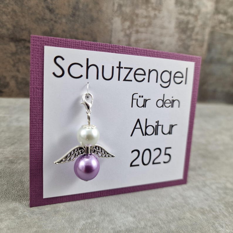 May include: A purple card with the text "Schutzengel f&uuml;r dein Abitur 2025" and a silver charm of an angel with white and purple beads.