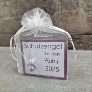 May include: A small purple and white angel charm with wings, hanging from a white organza bag. The bag has a card attached with the text "Schutzengel f&uuml;r dein Abitur 2025".