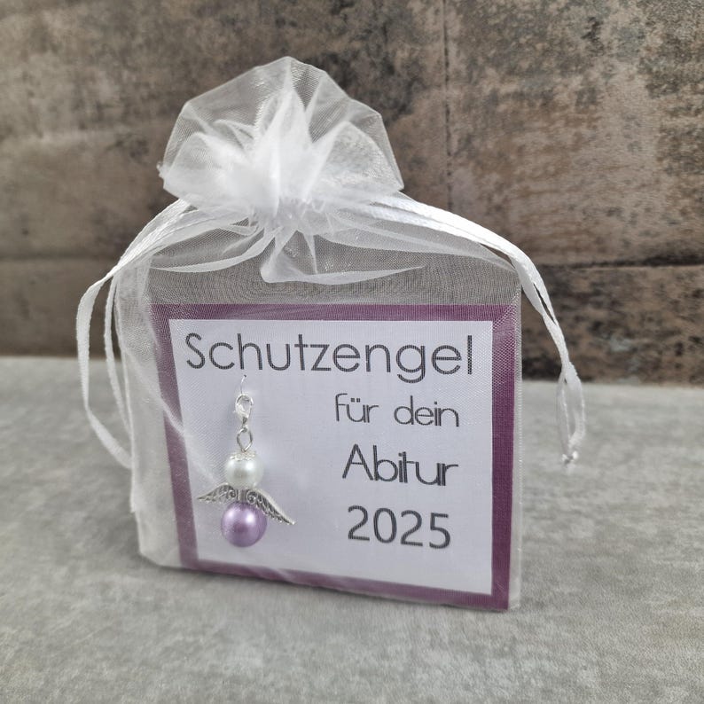 May include: A small purple and white angel charm with wings, hanging from a white organza bag. The bag has a card attached with the text "Schutzengel f&uuml;r dein Abitur 2025".