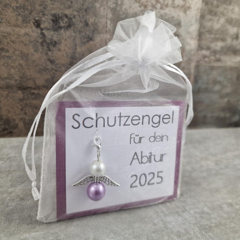 May include: A small purple and white angel charm with silver wings and a silver loop. The charm is in a white organza bag with a card that reads "Schutzengel f&uuml;r dein Abitur 2025".