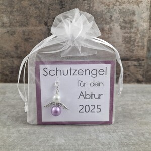 May include: A small white organza bag with a purple card that reads "Schutzengel f&uuml;r dein Abitur 2025". A silver charm with a purple bead and white beads hangs from the card.