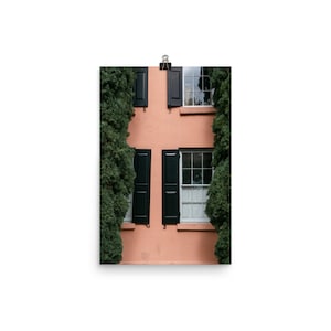 May include: A peach-colored building with two windows and black shutters. The windows have white trim and white curtains. Green bushes frame the building on either side.