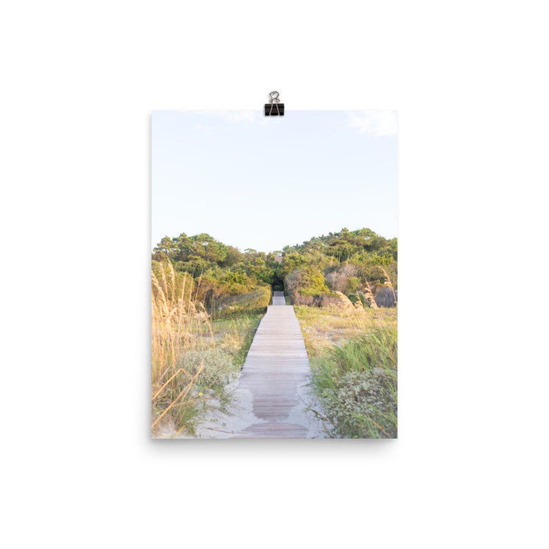 May include: A wooden walkway leads through a grassy area towards a line of trees in the distance. The sky is a light blue with some clouds.