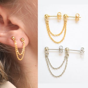 Könnte beinhalten: Goldene und silberne Doppelkettenohrringe. Die Ohrringe haben einen kleinen Kugelstecker, der mit zwei zarten Ketten verbunden ist. Die goldenen Ohrringe werden an einem Ohr getragen, die silbernen auf weißem Hintergrund.