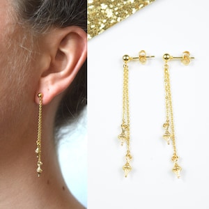 May include: Gold-coloured dangle earrings. Each earring has a small ball stud, a delicate chain, and three decorative charms. The earrings are shown on a white background with a gold glitter accent.