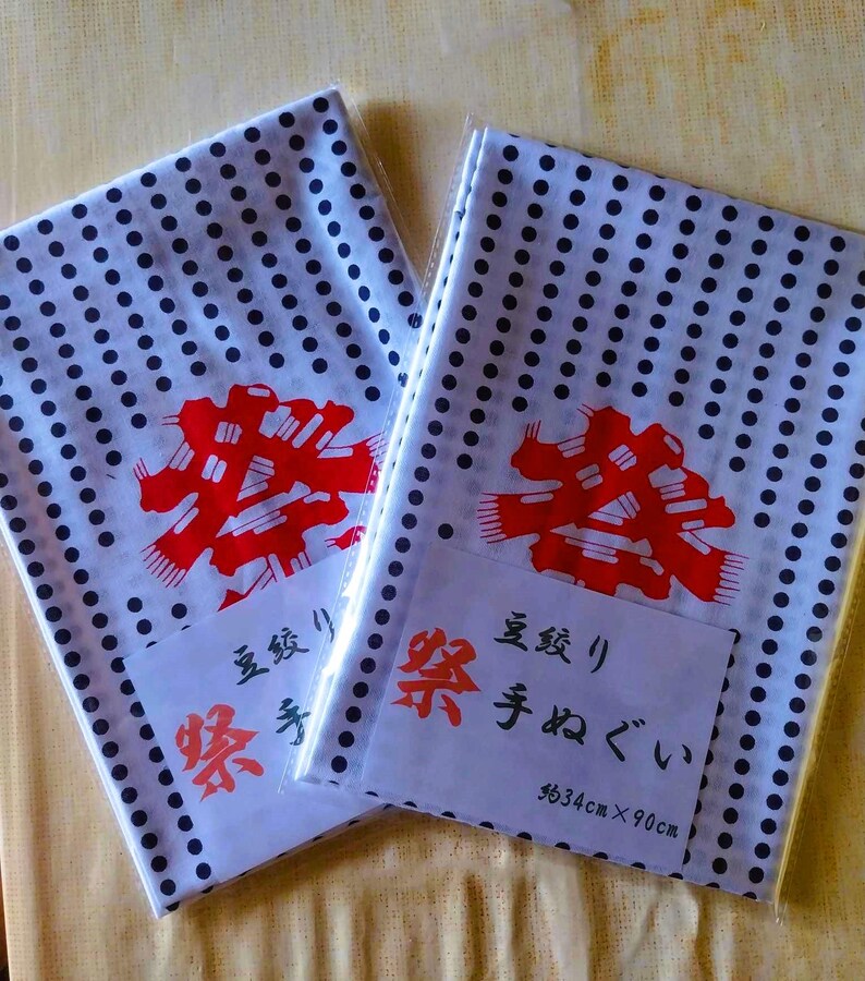 May include: Two white cotton handkerchiefs with a black polka dot pattern. Each handkerchief has a red kanji character printed on it. The handkerchiefs are packaged with a white label that reads "長さ" and "手ぬぐい" in Japanese. The label also includes the dimensions "approx 34cm x 90cm".