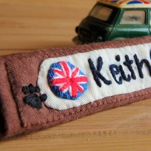 Peut inclure: Une étiquette de nom en tissu marron avec le nom "Keith" brodé en noir. L'étiquette comporte deux drapeaux Union Jack brodés et une empreinte de patte noire. Une voiture jouet verte est en arrière-plan.