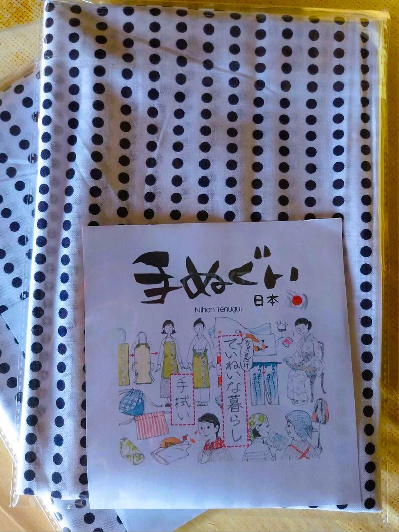 May include: A white fabric with black polka dots and a Japanese tenugui with illustrations of different ways to wear the tenugui. The tenugui has the text "手拭い" which means "hand towel" in Japanese. The text "てぬぐい" is also written in Japanese. The text "Nihon Tenugui" is written in English. The text "日本" is written in Japanese and means "Japan".