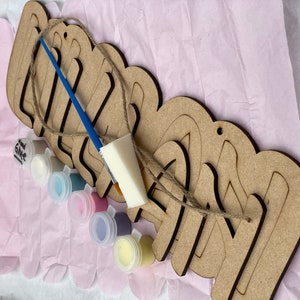 May include: A wooden sign with the word "Olivia" spelled out in large letters. The sign is unfinished and comes with a set of six paint pots in various colors, a paintbrush, and a sponge. The sign is ready to be painted and personalized.