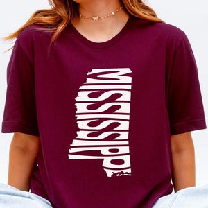 May include: A maroon t-shirt featuring the state of Mississippi in white, with the state's name vertically stacked within the outline. The shirt has short sleeves and a crew neck. The person is wearing light blue jeans.