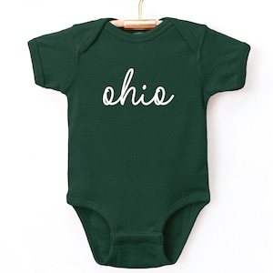 May include: A dark green baby onesie with the word "ohio" in white script. The onesie is hanging on a gold hanger from a wooden rack. The background is white.