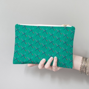May include: A teal zippered pouch with a repeating pattern of crossed lines and dots. The pouch is being held by a hand.