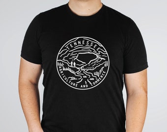 May include: A black short-sleeved t-shirt featuring a white circular graphic on the chest. The design depicts a landscape with a river and mountains, surrounded by the text TENNESSEE AGRICULTURE AND COMMERCE in a bold, clean, sans-serif font.