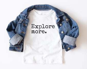 May include: A white t-shirt with the words "Explore more." in black, paired with a blue denim jacket. The jacket is draped over the shirt, with the sleeves rolled up. The overall aesthetic is casual and youthful.