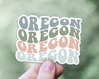 May include: A sticker with the word "OREGON" in a retro font, layered in shades of blue, green, and peach. The sticker has a white border and is held against a blurred green background.