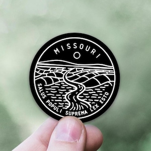 May include: A black round sticker featuring a white illustration of a river winding through a landscape. The word "MISSOURI" arches across the top, with "SALUS POPULI SUPREMA LEX ESTO" around the bottom.