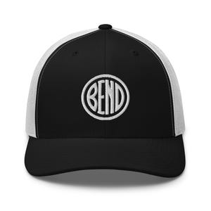 May include: Black and white trucker hat with a curved brim. The front panel is black with a white embroidered circular design that reads "BEND". The back of the hat is white mesh.