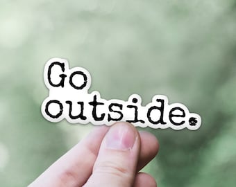 May include: A white sticker with black text that reads "Go outside." The sticker is being held by a hand against a green blurred background.