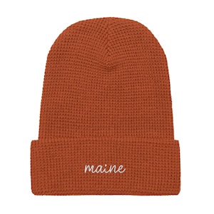 May include: A rust-colored beanie hat with a waffle knit texture. The word "maine" is embroidered in white script on the folded brim. The hat is a warm, casual accessory.