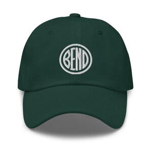 May include: A dark green baseball cap with a curved brim. The cap features the white embroidered word "BEND" inside a white circular design on the front. The hat is made of a soft fabric and has a classic, adjustable fit.