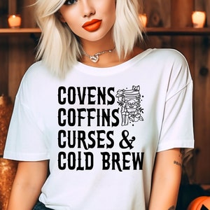 以下が含まれることがあります： 黒い文字で「Covens Coffins Curses & Cold Brew」と書かれた白いTシャツ。テキストは、ドクロとクロスボーンのグラフィックと花のアクセントでスタイリッシュにデザインされています。