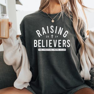 Puede incluir: Una camiseta gris oscuro con el texto "Raising Believers" en blanco, con una cruz y los números 20 y 24 debajo del texto. El texto "The Praying Moms Club" está debajo de los números.
