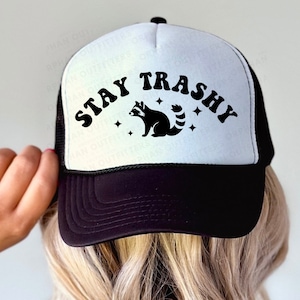 May include: A black and white trucker hat with a white front panel and a black mesh back. The front panel features the text "STAY TRASHY" in a retro font with a black raccoon illustration and stars.